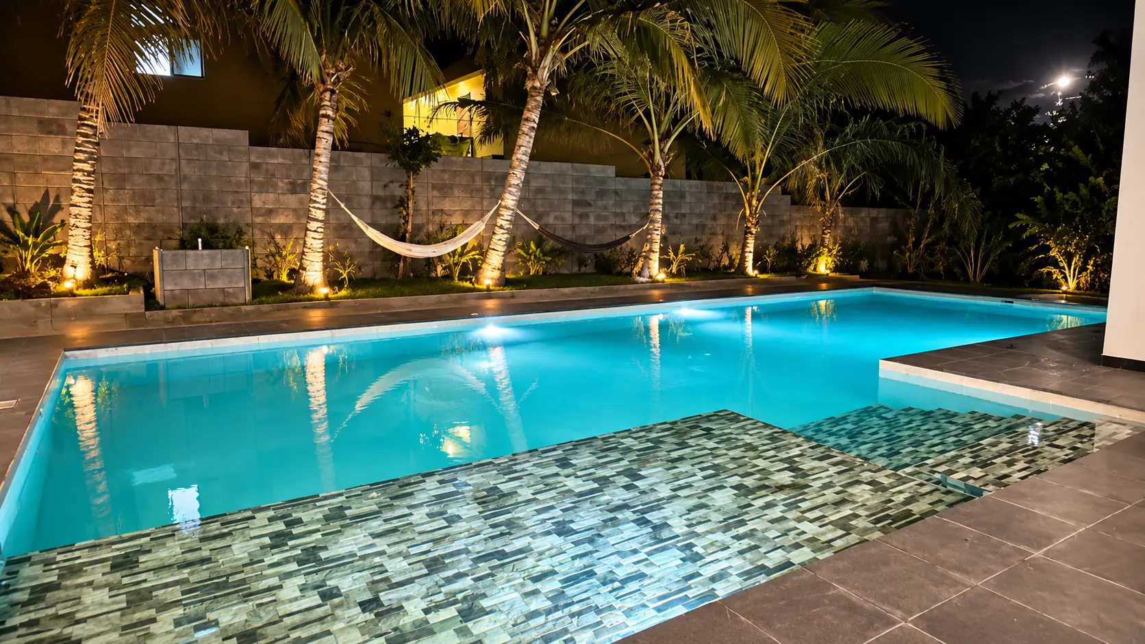 Piscina iluminada de noche entre palmeras y hamaca